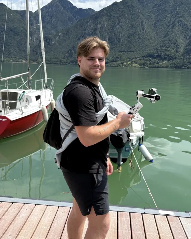 Gianni Adam bij een meer, met een zeilboot en bergen op de achtergrond